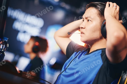 Preview: Side view of a confused asian guy, male cyber sport gamer wearing headphones looking at