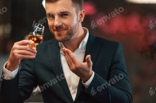 Preview: Positive elegant man in black suit is in the bar, tasting the alcohol drink and smiling