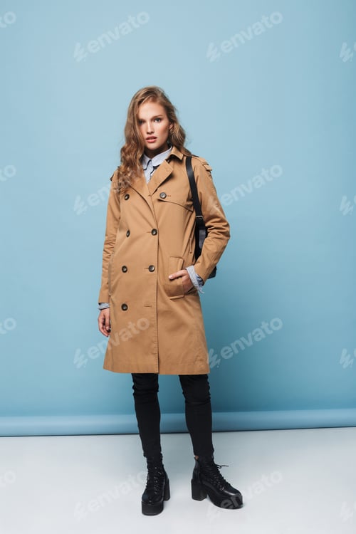 Preview: Pensive model with wavy hair in trench coat with backpack intently looking in camera at studio