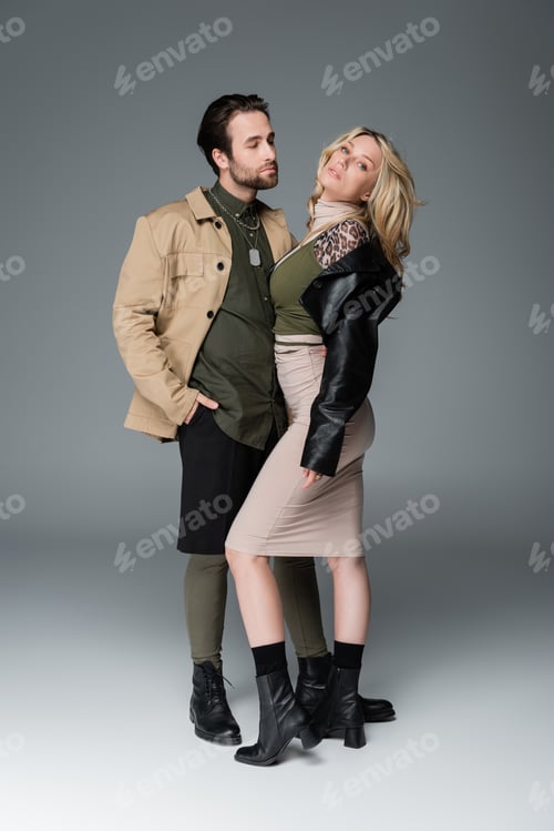 Preview: full length of stylish man in autumnal outfit posing with hand in pocket near blonde woman on grey