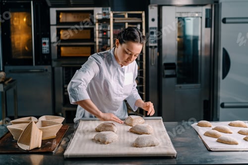 Preview: concentrated beautiful woman baker forms raw bread buns before baking bakery production of pastries