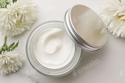 Preview: Glass jar of face cream and flowers on white marble table, flat lay