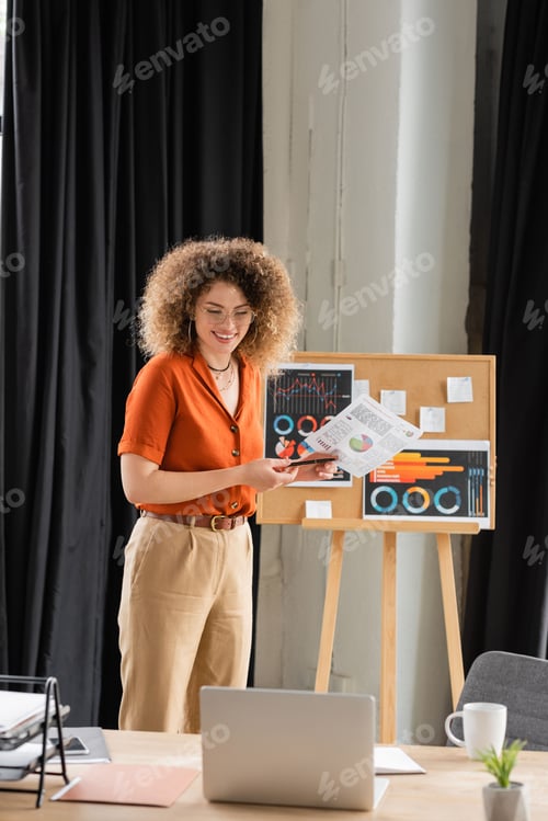 Preview: happy businesswoman in glasses holding infographics near office board during video call