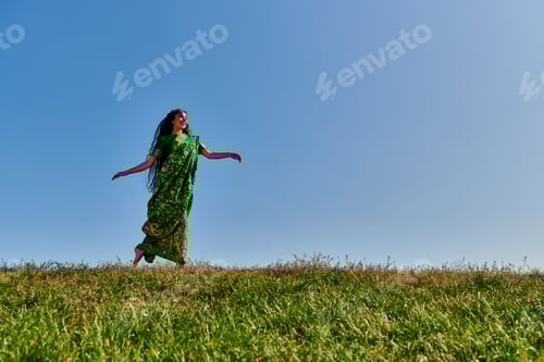 Preview: vibrant summer, joyful indian woman in ethnic clothes running in green field under blue sky