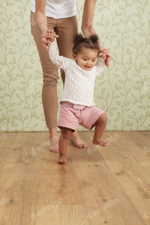 Preview: Mother holding baby girl taking first steps, low section