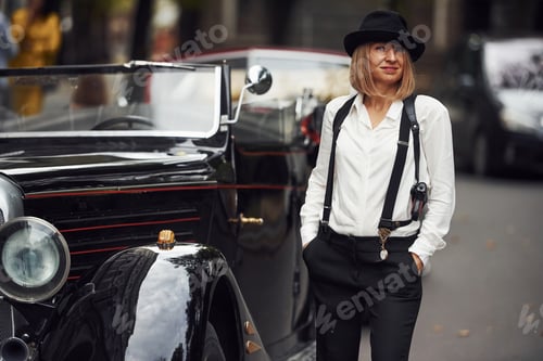 Preview: Blonde woman in hat and in black retro clothes near old vintage classic car