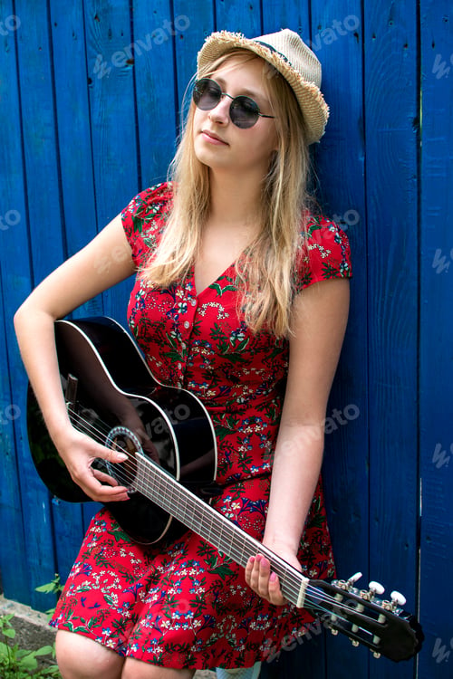 Preview: Beautiful teenage blonde long haired girl playing acoustic guitar outdoor over blue wooden fence