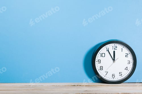 Preview: Clock showing five to twelve deadline coming soon on wood office desk with blue wall