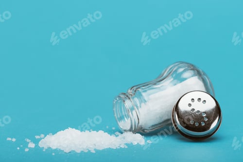 Preview: open salt shaker and metallic cap near sprinkled salt on blue background with copy space