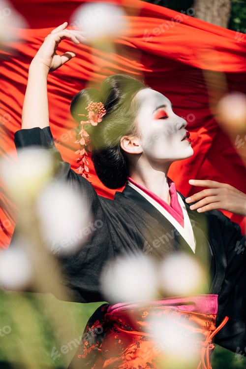 Preview: selective focus of tree branches and beautiful geisha dancing with red cloth on background in