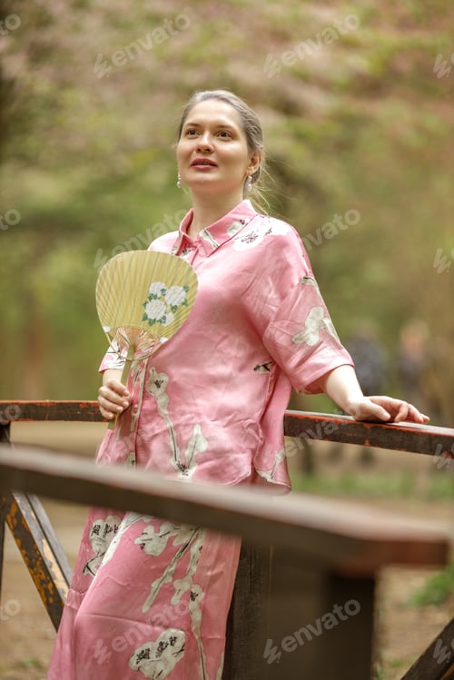 Preview: Young woman in pink national costume of Japan, walking in a garden with blooming sakura