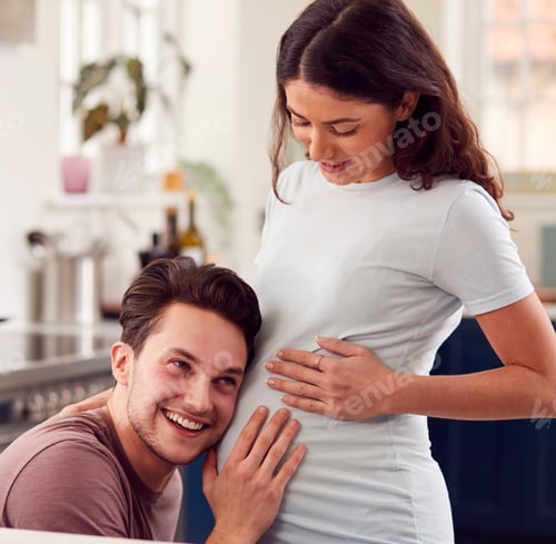Preview: Excited Pregnant Transgender Couple At Home In Kitchen With Man Listening To Baby's Heartbeat