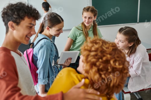 Preview: Cheerful teenage pupils talking and using digital tablet near blurred friends in classroom in school