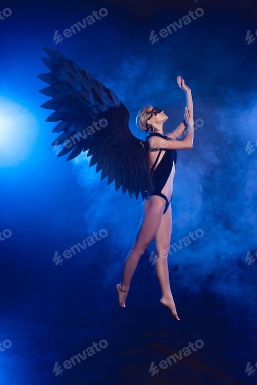 Preview: beautiful sexy woman with lace mask and black angel wings jumping on dark blue background
