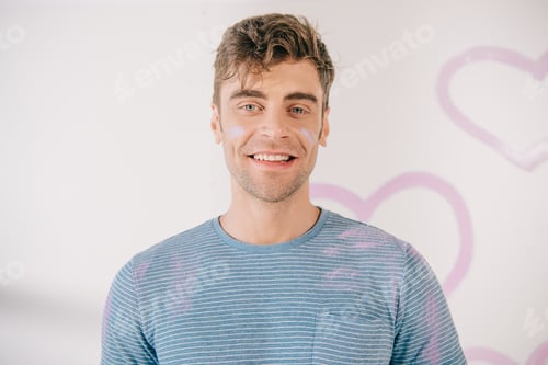 Preview: handsome young man with stains of pink paint on face looking at camera