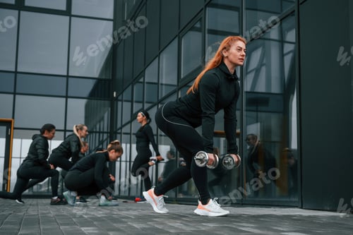 Preview: Lifting dumbbells. Group of sportive women is outdoors near black building