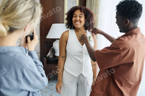 Preview: Happy young African American bride posing in front of bridesmaid with smartphone