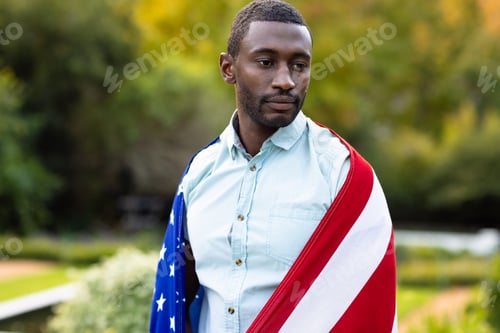 Preview: Serious african american man covered with flag of usa in garden