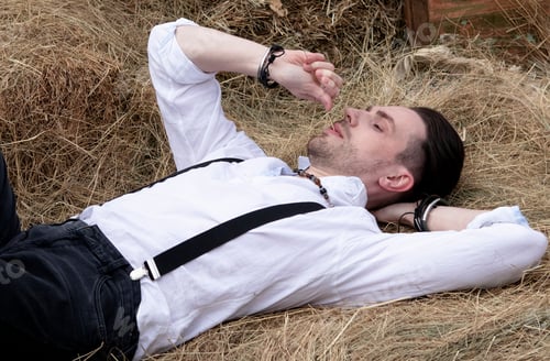 Preview: the groom in a white shirt and black hat lies on a haystack. Rural wedding