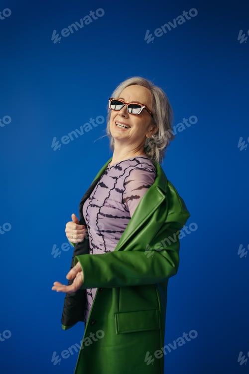 Preview: senior woman in sunglasses and green leather jacket smiling isolated on blue