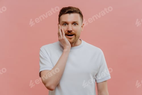 Preview: Amazed bearded man keeps hand on cheek stands speechless cannot believe his eyes
