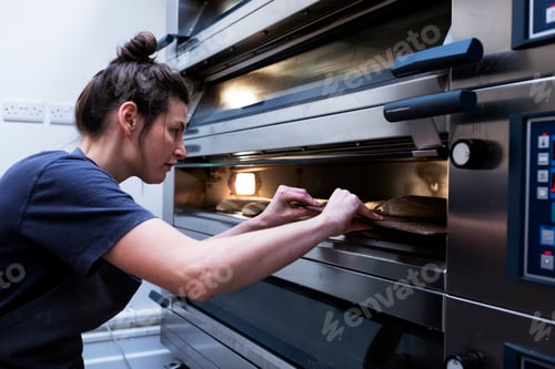 Preview: Woman wearing apron standing in an artisan bakery, placing sourdough breads into oven.