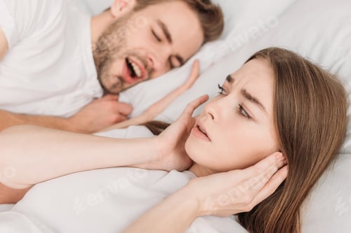 Preview: selective focus of woman plugging ears with hands while lying in bed near snoring husband
