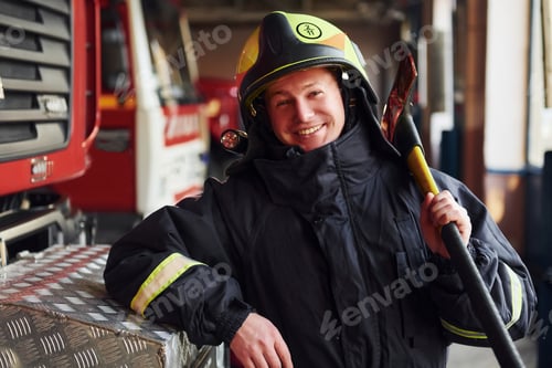 Preview: Holds axe in hands. Male firefighter in protective uniform standing near truck