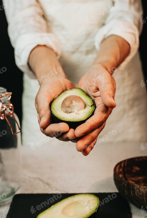 Preview: Woman chef holding and showing ripe cut avocado on white kitchen apron.Half avocado on black board,w