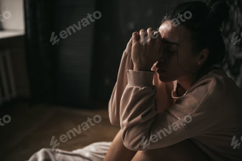 Preview: Candid sad brunette with bun of hair in beige clothes of earth tones girl sit on the bed and suffers