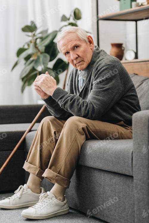 Preview: upset retired man sitting on sofa near walking stick in living room