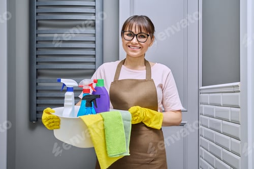 Preview: Middle-aged woman in gloves apron with basin of detergents in bathroom