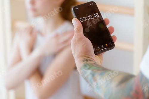 Preview: Coach holding a phone with a stopwatch, counting the time of a young girl training at the gym