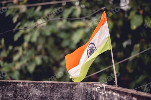 Preview: Closeup shot of the Indian flag hung on the edge of a balcony with green leaves in the background