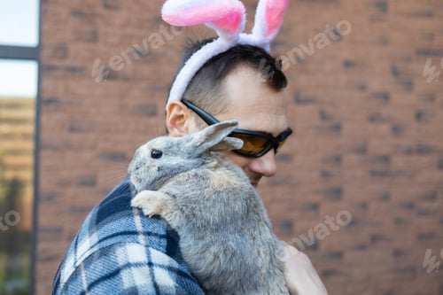 Preview: Portrait of happy funny handsome young man in bunny ears standing near house, looking at camera, hol