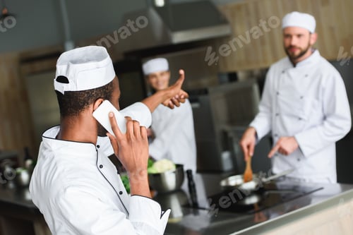 Preview: african american chef talking by smartphone and showing thumb up to colleagues at restaurant kitchen