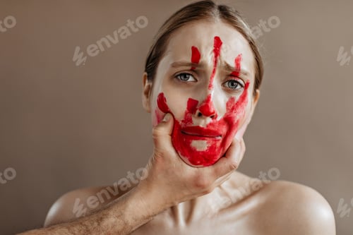 Preview: Unhappy woman with bloody hand print on her face is roughly taken by throat