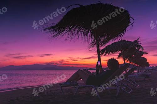 Preview: A woman photographing the sunset sitting on a relaxing beach chair with a parasol at night