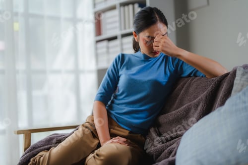 Preview: Stressed asian woman pressing forehead, suffering intense migraine while sitting on living room sofa