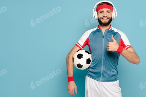 Preview: happy stylish sportsman in headphones with soccer ball showing thumb up on blue background