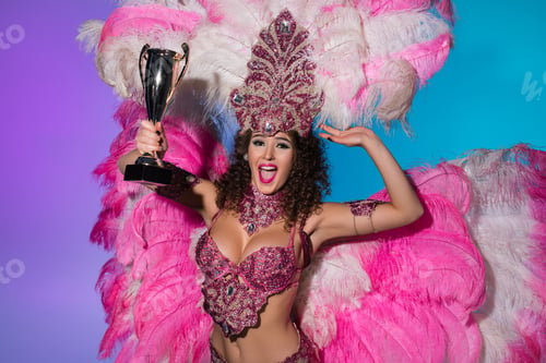 Preview: Happy young woman in carnival costume with pink feathers holding winner cup isolated on blue