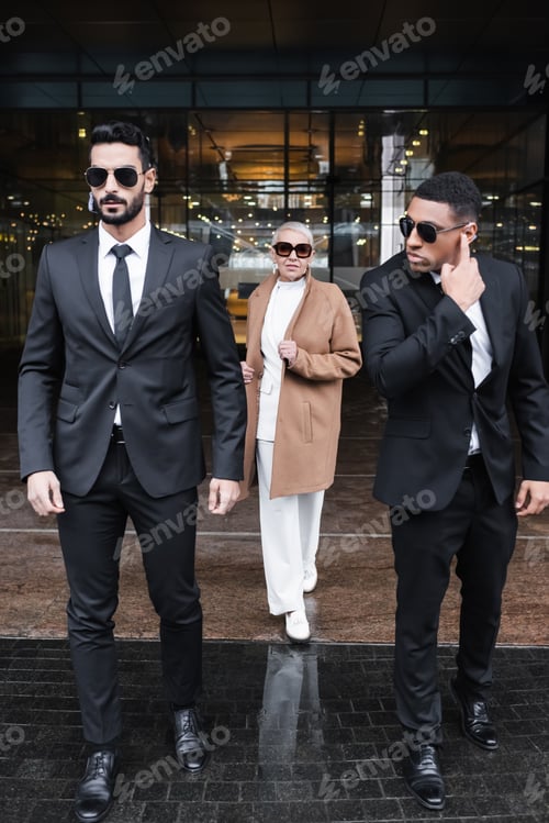 Preview: interracial bodyguards in sunglasses escorting stylish businesswoman near hotel building