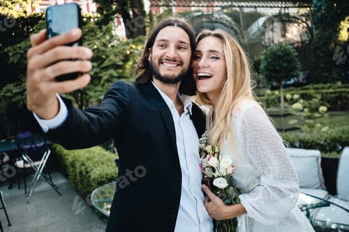 Preview: Young attractive groom and bride joyfully taking selfie together in beautiful garden of restaurant