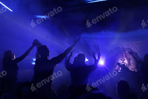 Preview: People Raising hands against blue smoky background with light beams during concert and crowd of peop