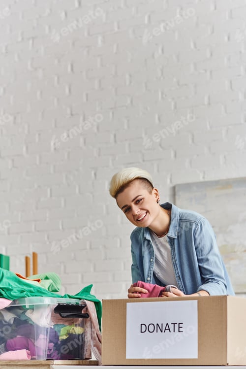 Preview: young and joyful woman with trendy hairstyle holding pre-loved clothes and looking at camera