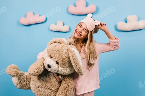 Preview: Romantic girl posing with toy rabbit. Studio shot of cheerful woman in pink pajama standing on blue