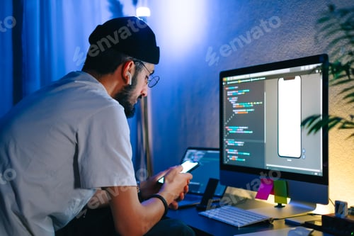 Preview: Young man mobile developer writes program code on a computer and check app in smartphone