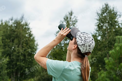 Preview: Young blonde woman bird watcher in cap looking through binoculars at cloudy sky in summer forest
