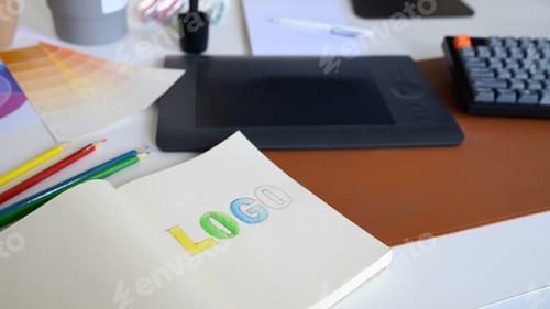 Preview: Close-up of logo sketch with color pencils, palette, and tablet on designer workspace