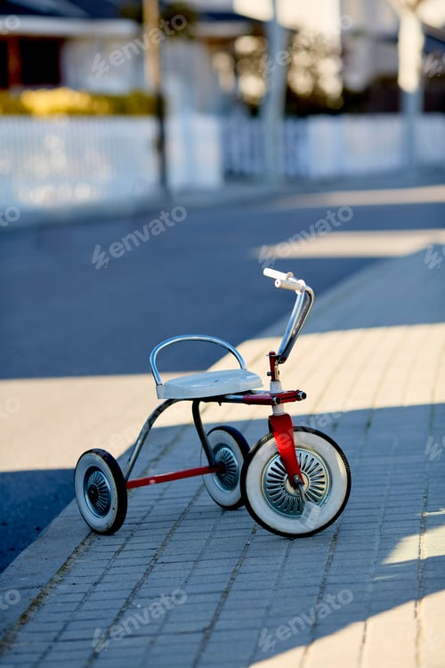 Preview: A child's red and white tricycle is parked on a sunlit sidewalk, casting a soft shadow.